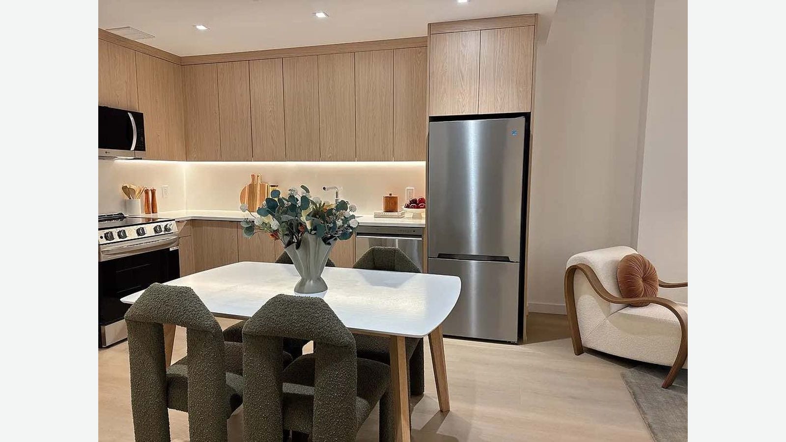 Photograph of an open kitchen with pale wood cabinets, a white counter, a stainless-steel refrigerator, a smooth-top range, a microwave over it, and a white dining table with gray upholstered chairs in front