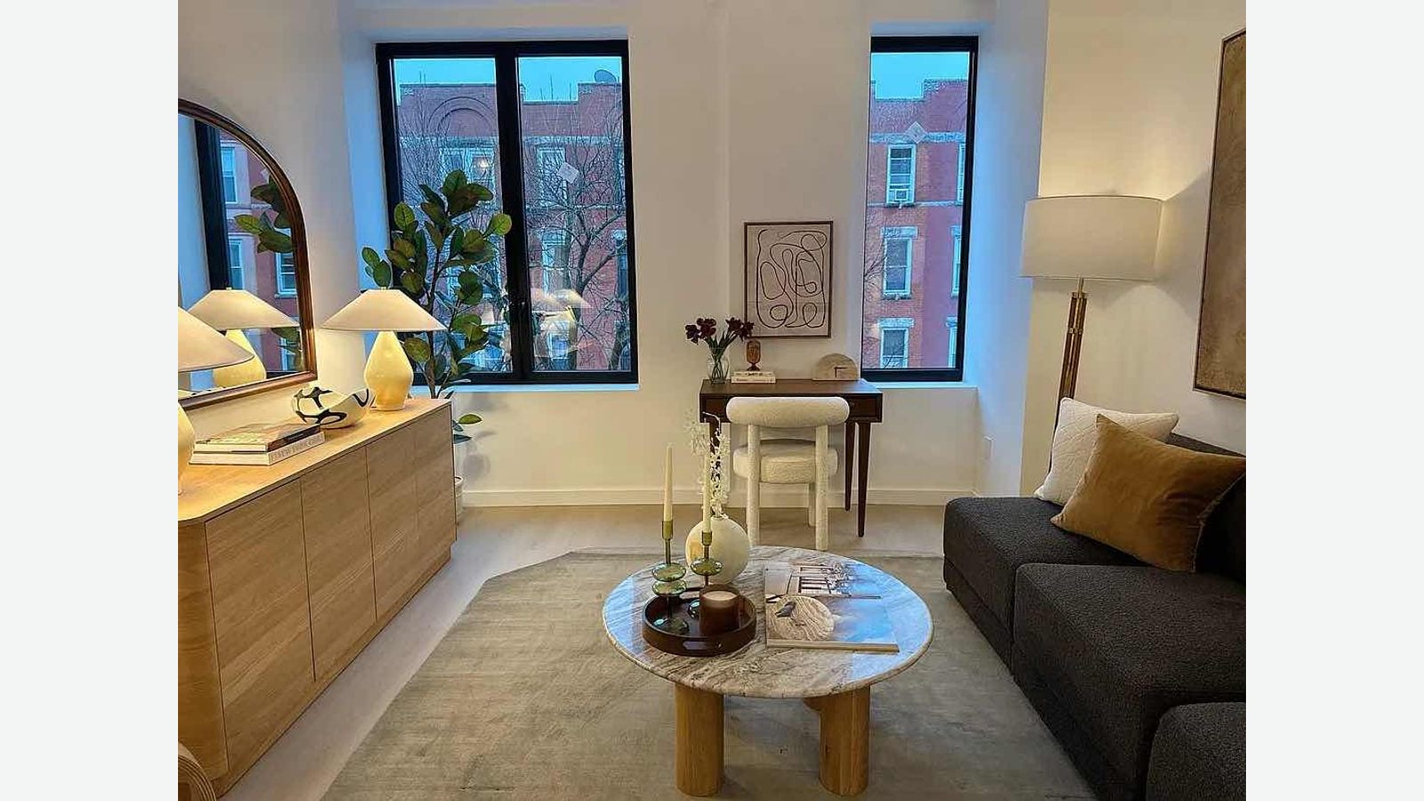Photograph of a furnished living room: two tall black-framed windows looking onto red-brick row houses, a dark gray sofa, a round marble coffee table, a wood sideboard with two lamps, an arched mirror and a small desk