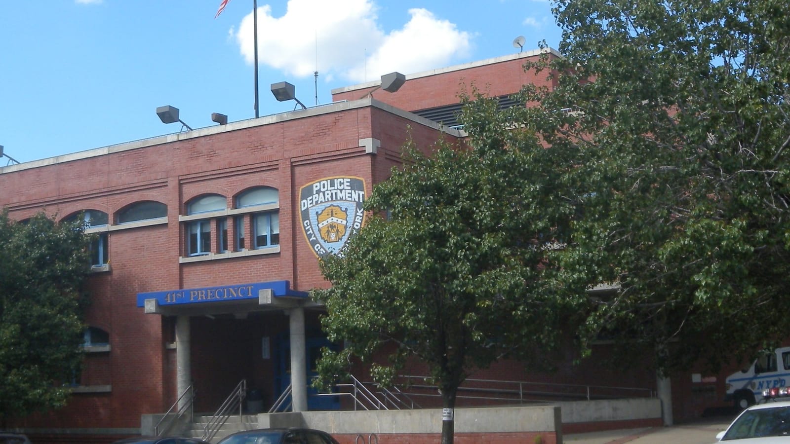Photograph of a two-story red brick police station with arched windows, a Police Department City of New York shield on the wall, a blue canopy reading 41st Precinct over the entrance steps, a leafy tree in front and parked cars, including a police car, along the curb