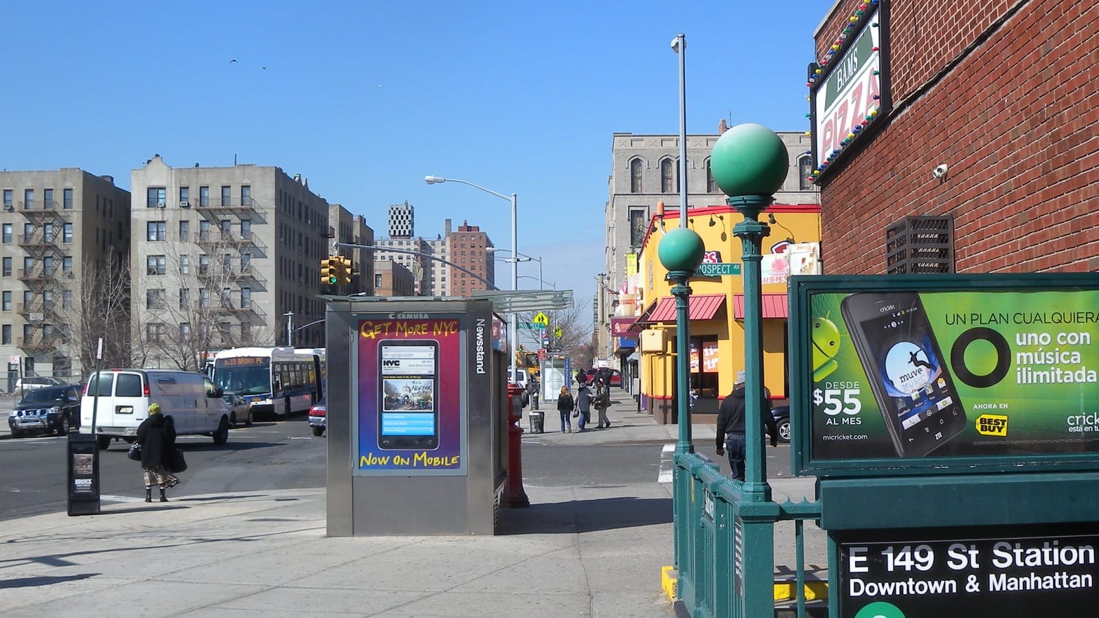 Photograph of a green iron subway stair entrance with globe lamps and a sign reading E 149 St Station, Downtown and Manhattan, 6, beside a brick wall and a phone advertisement, with a steel newsstand, a city bus, parked vans and six-story brick apartment buildings along a wide street under a blue sky