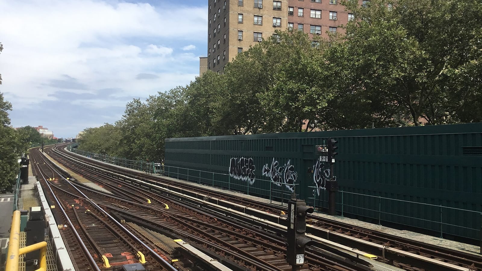 Photograph from a raised subway platform looking along several curving tracks with switches and a signal, a dark green wall with graffiti at right, a row of leafy trees and a tall brick apartment tower behind under a partly cloudy sky