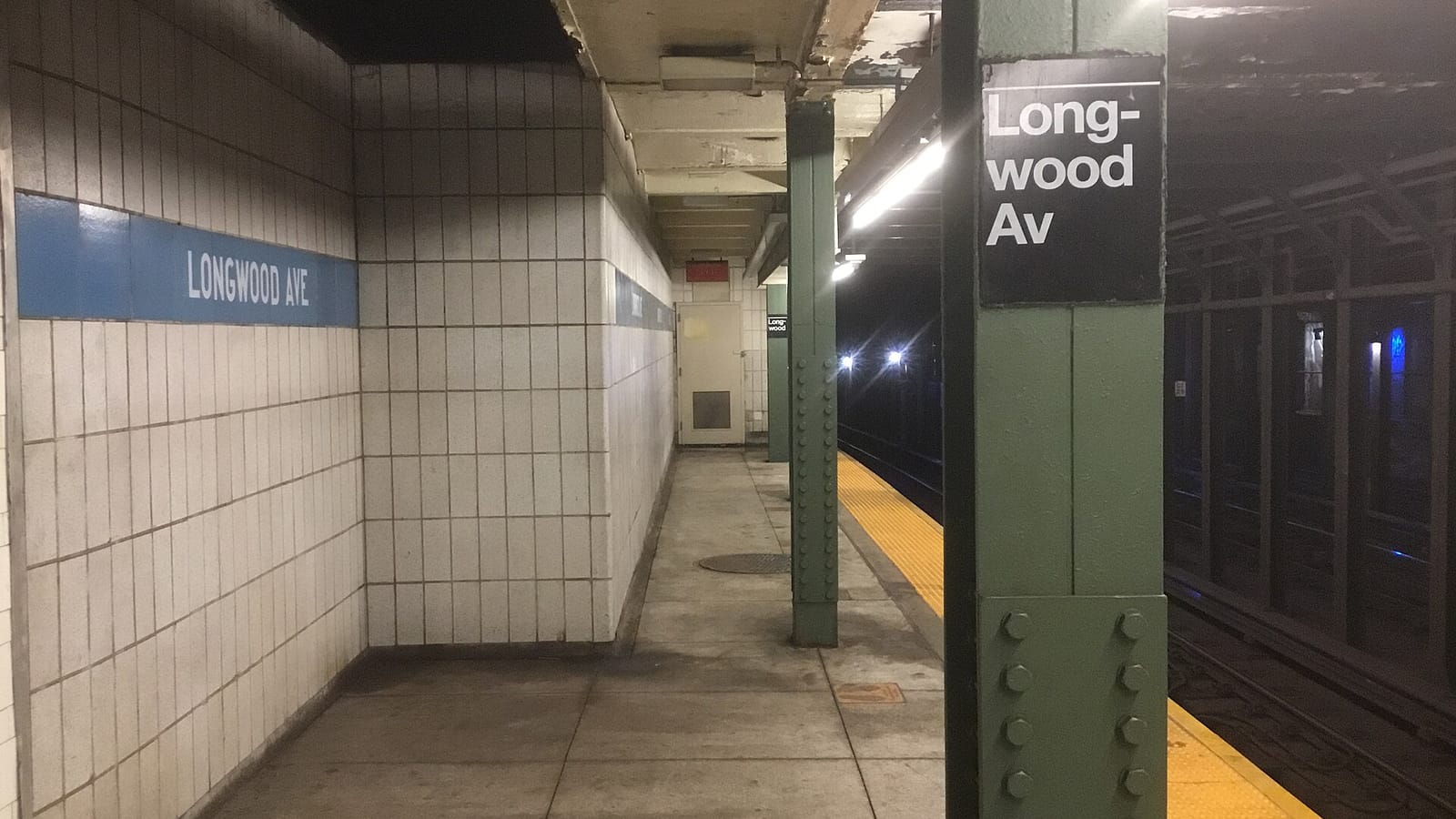 Photograph of an empty underground subway platform with white tiled walls, a blue band reading Longwood Ave, green steel columns, a black sign reading Longwood Av and a yellow platform edge beside the tracks