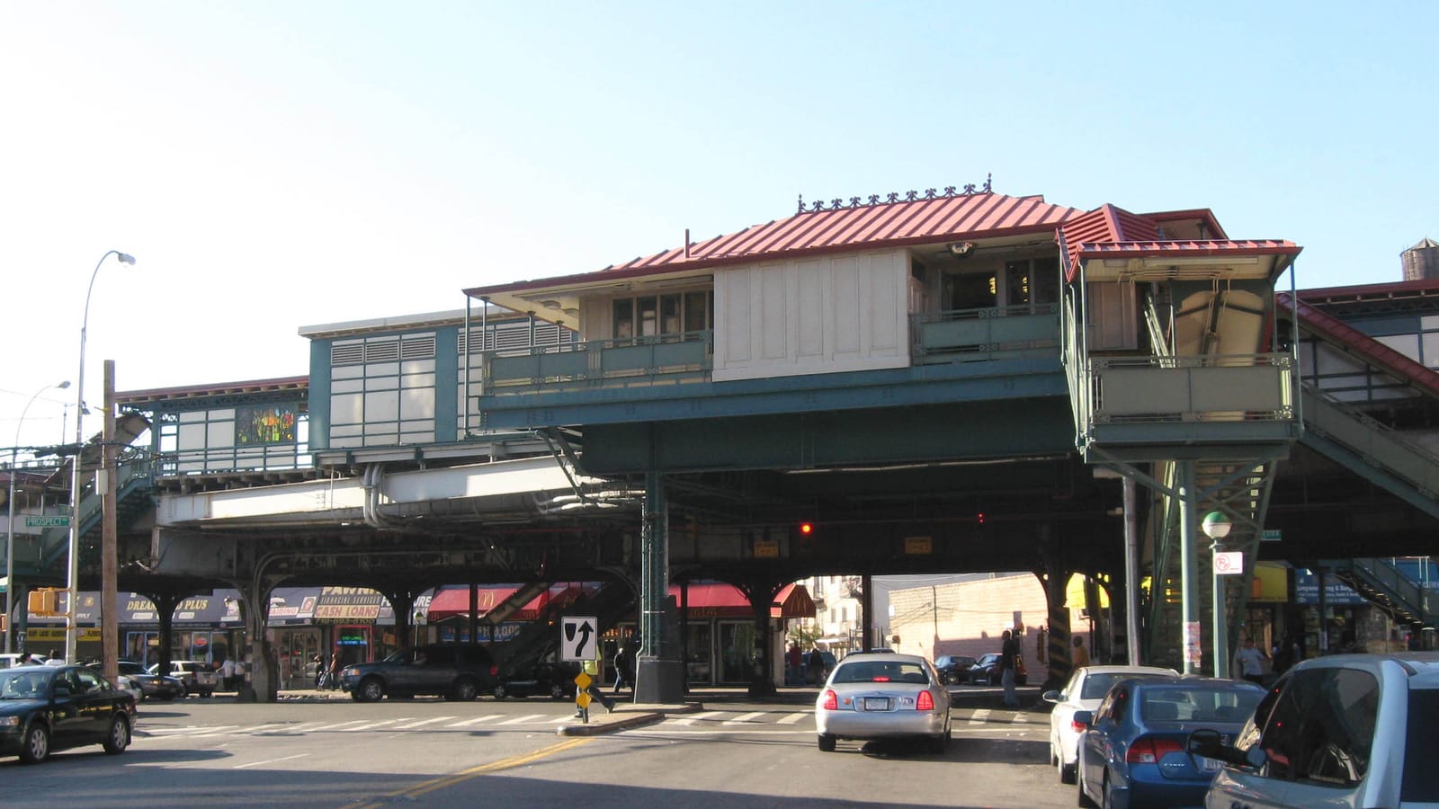 Photograph of a raised subway station with a red metal roof, pale green steel supports and stairways at both ends, over a wide street with cars, a crosswalk and storefronts with awnings underneath, on a clear afternoon