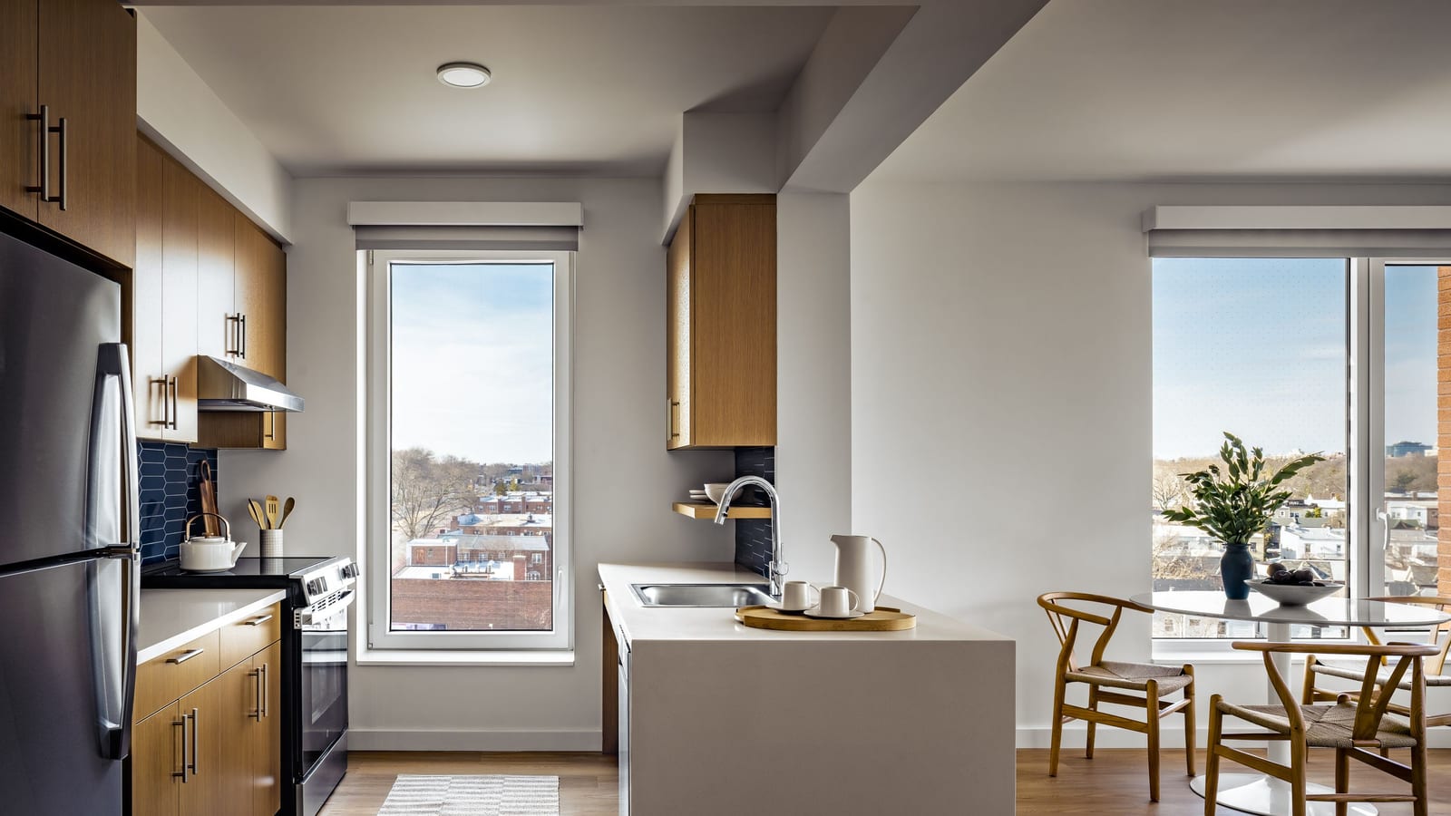Photograph of a kitchen with wood-tone cabinets, a stainless-steel refrigerator, a smooth-top electric range under a hood, a dark blue tile backsplash, a white peninsula with a sink, tall windows looking over low brick houses and a small round table with wooden chairs