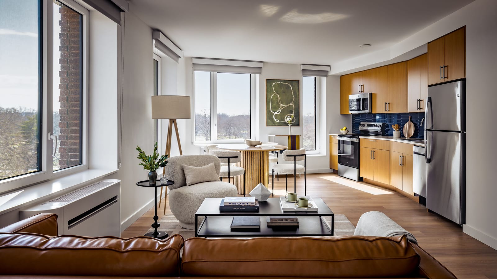 Photograph of a furnished corner living room with windows on two walls looking over treetops, a brown leather sofa, a round dining table with white chairs, wood-look plank flooring, a white unit under the window at the left and, at the right, a kitchen with wood-tone cabinets, a dark blue tile backsplash, a microwave, an electric range, a dishwasher and a stainless-steel refrigerator