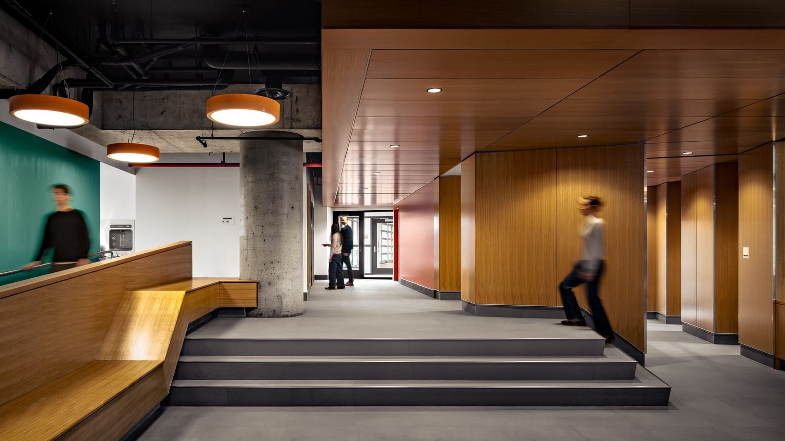Photograph of a lobby with three wide grey steps, a built-in wooden bench along a low wall, a round concrete column, orange drum-shaped ceiling lights under exposed concrete and pipes, wood-paneled walls and ceiling at the right, a teal wall at the left and four blurred figures walking or standing