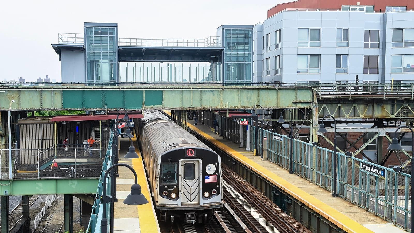 Photograph from above of an elevated station: an L train with its headlights on pulls into the platform, a glass-walled elevator tower and enclosed overpass stand over the tracks, a sign on the right-hand fence reads Livonia Avenue with the accessibility symbol, and a modern apartment building rises at the upper right