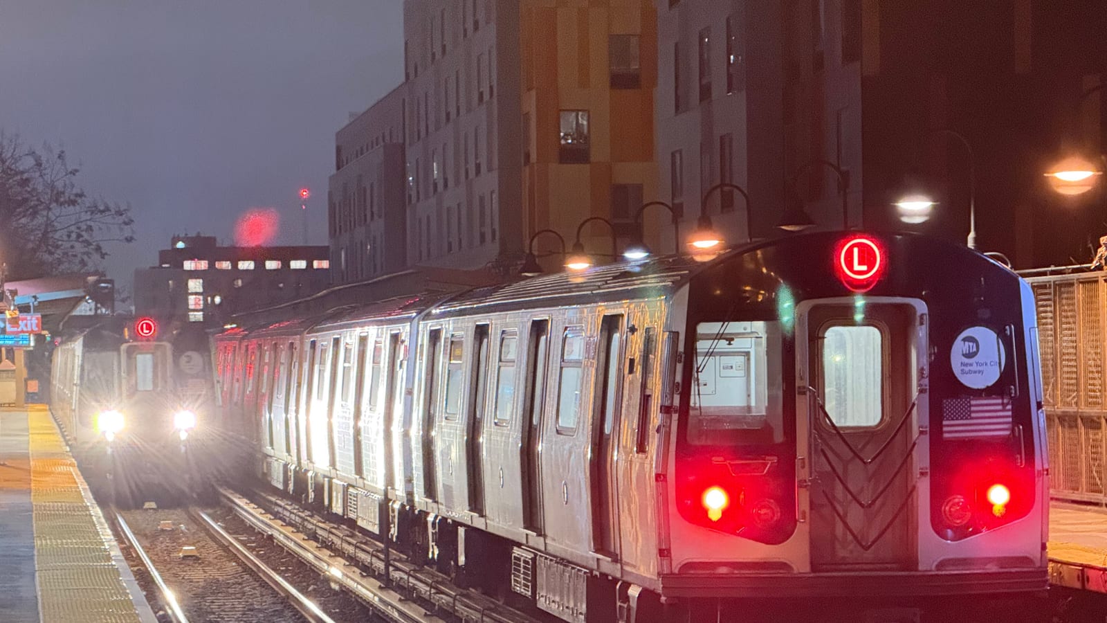 Night photograph of two silver L trains at an elevated station platform: the nearer train shows its rear with red tail lights, a red L route sign and an MTA New York City Subway decal, the second approaches with bright headlights, and tall apartment buildings with lit windows stand behind the platform lamps