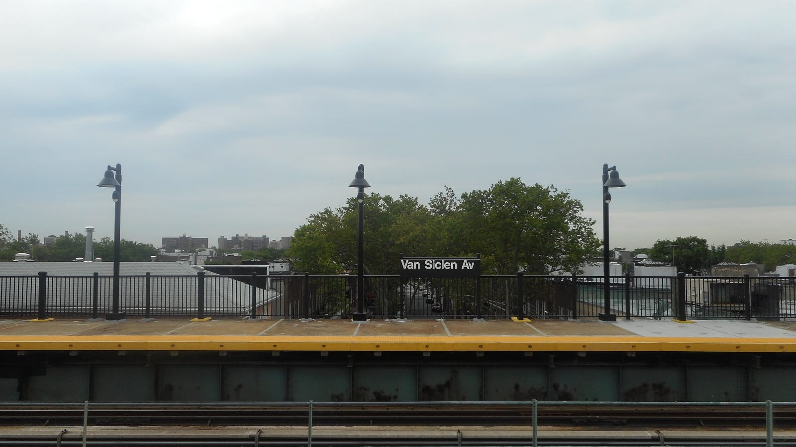 Photograph across elevated subway tracks to a station platform with a yellow edge strip, black railings and three lamp posts, a black sign reading Van Siclen Av in the middle, and low rooftops and trees under a grey sky behind