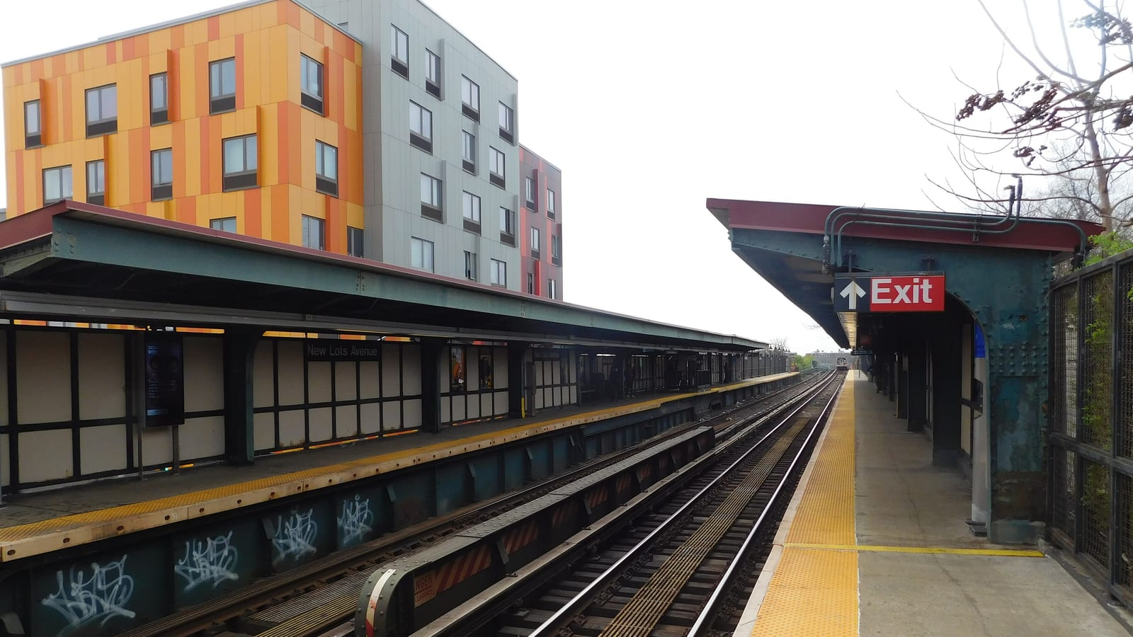 Photograph of an elevated subway station: two wooden-walled platforms with green steel canopies on either side of the tracks, a sign reading New Lots Avenue on the left platform wall, a red Exit sign over the right platform, yellow tactile strips along both edges and a five-story apartment building with orange and grey panels behind the left platform