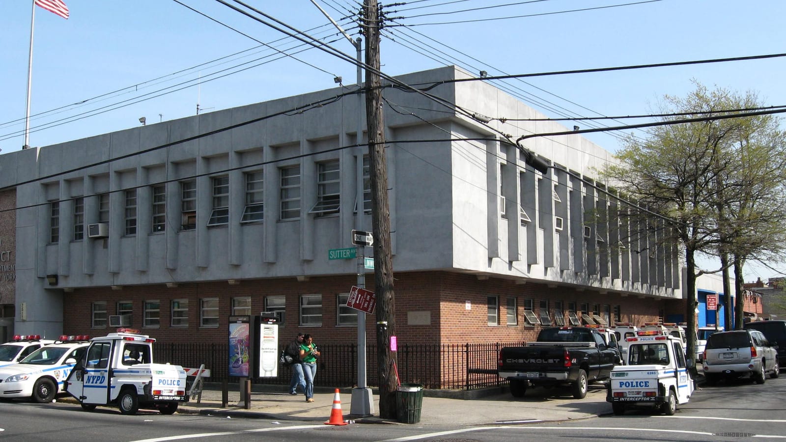 Photograph of a two-story police station with a gray concrete upper floor and a red brick ground floor behind a black iron fence, at a street corner with green signs reading Sutter Av and Linwood, NYPD cars and small three-wheeled police vehicles parked along the curb, a wooden utility pole and an American flag on a pole at left