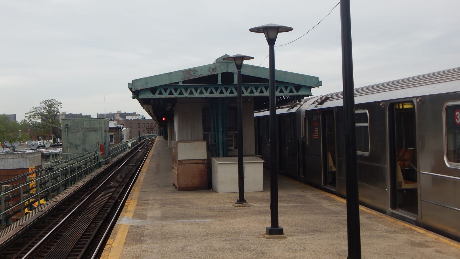 Photograph of an elevated subway platform with a green-painted steel canopy, black lamp posts, two metal cabinets and a silver train with a red 3 sign at right, empty tracks at left, and rooftops and tall apartment buildings in the distance under a gray sky