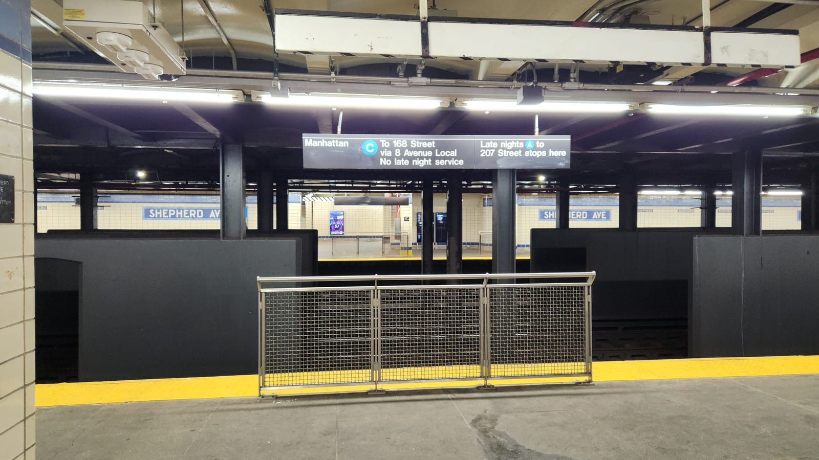 Photograph of an underground subway platform with a yellow edge strip and a short metal mesh barrier, a hanging sign reading Manhattan, C to 168 Street via 8 Avenue Local, no late night service, and late nights A to 207 Street stops here, with the opposite platform and Shepherd Ave name tiles on the far wall