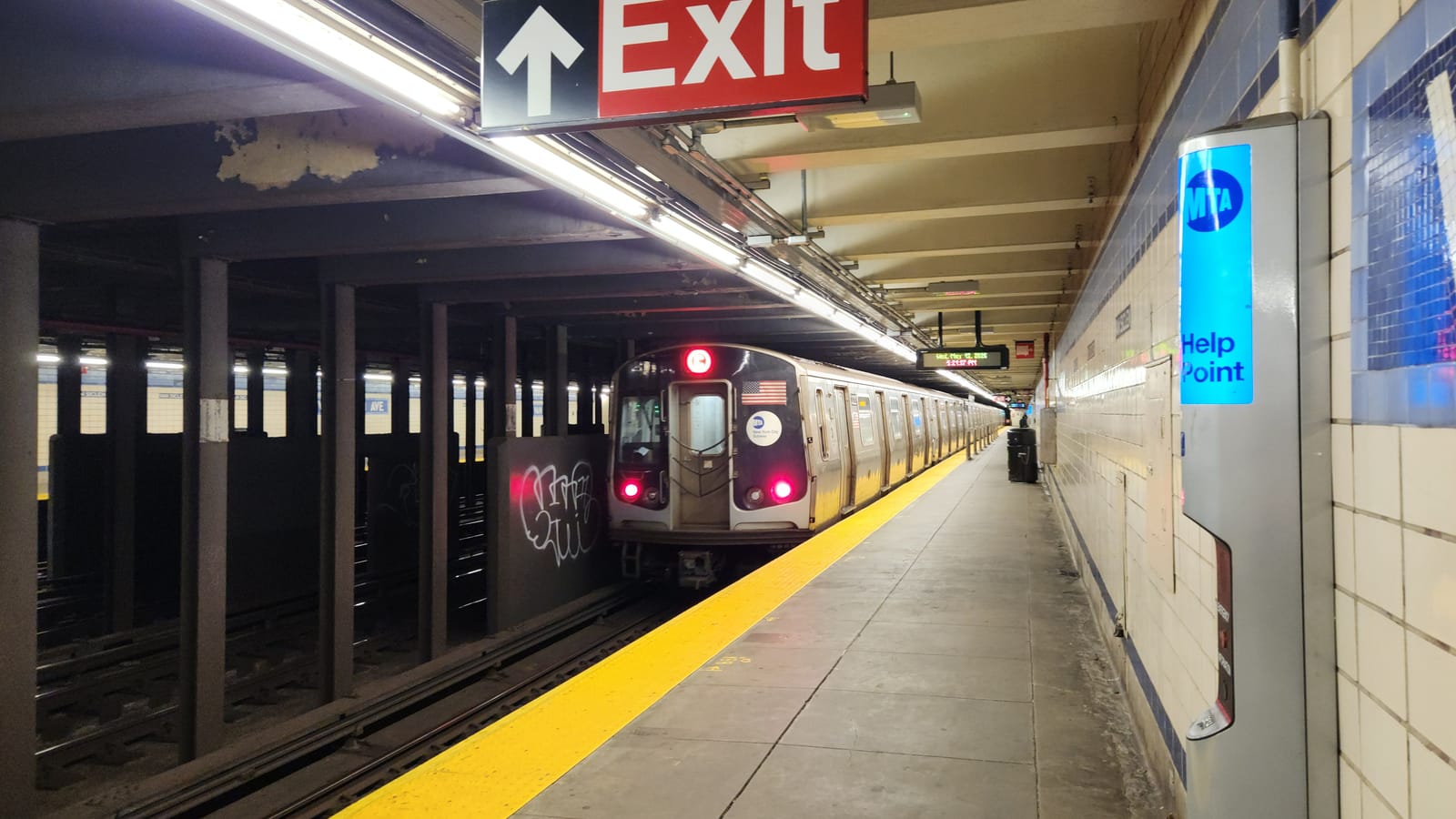 Photograph of an underground subway platform with a yellow edge strip, a silver train with red lights and an American flag decal pulling away, a red Exit sign overhead, a blue MTA Help Point on the tiled wall at right, and dark steel columns at left