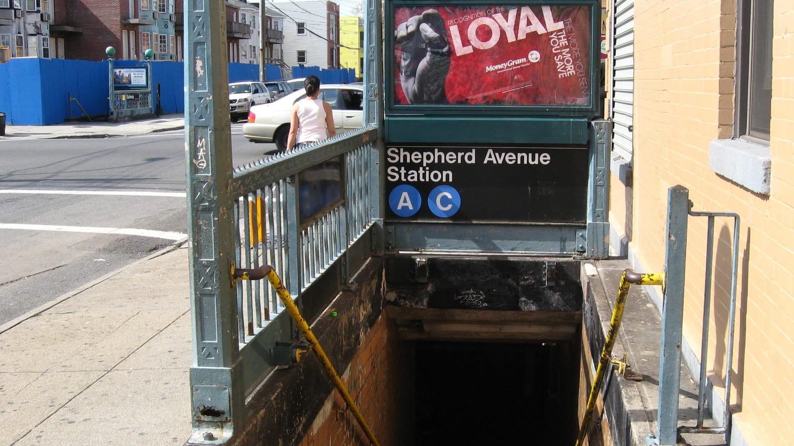 Photograph of a subway stair going down from a sidewalk, under a green iron railing with a black sign reading Shepherd Avenue Station and blue circles marked A and C, a red print advertisement above the sign, a tan brick wall at right, and a street with a white car, a woman walking away and blue construction fencing behind