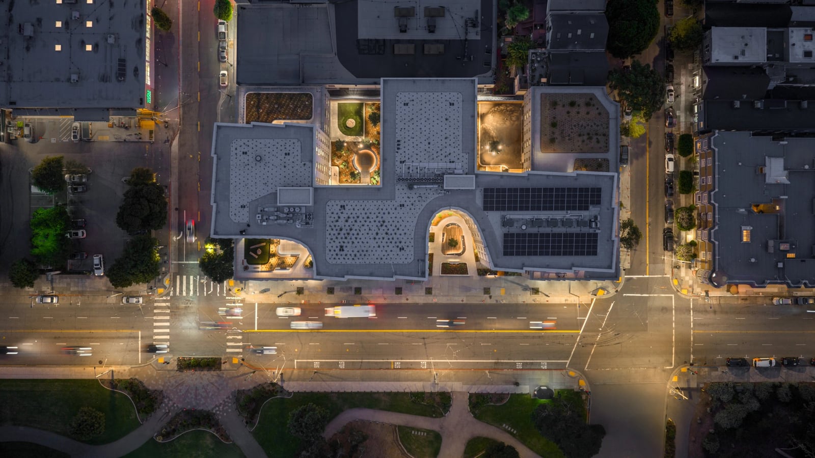 Aerial view straight down on a building at dusk: gray roofs with solar panels, three lit courtyards cut into the block, a wide street with moving traffic below and park paths at the bottom edge