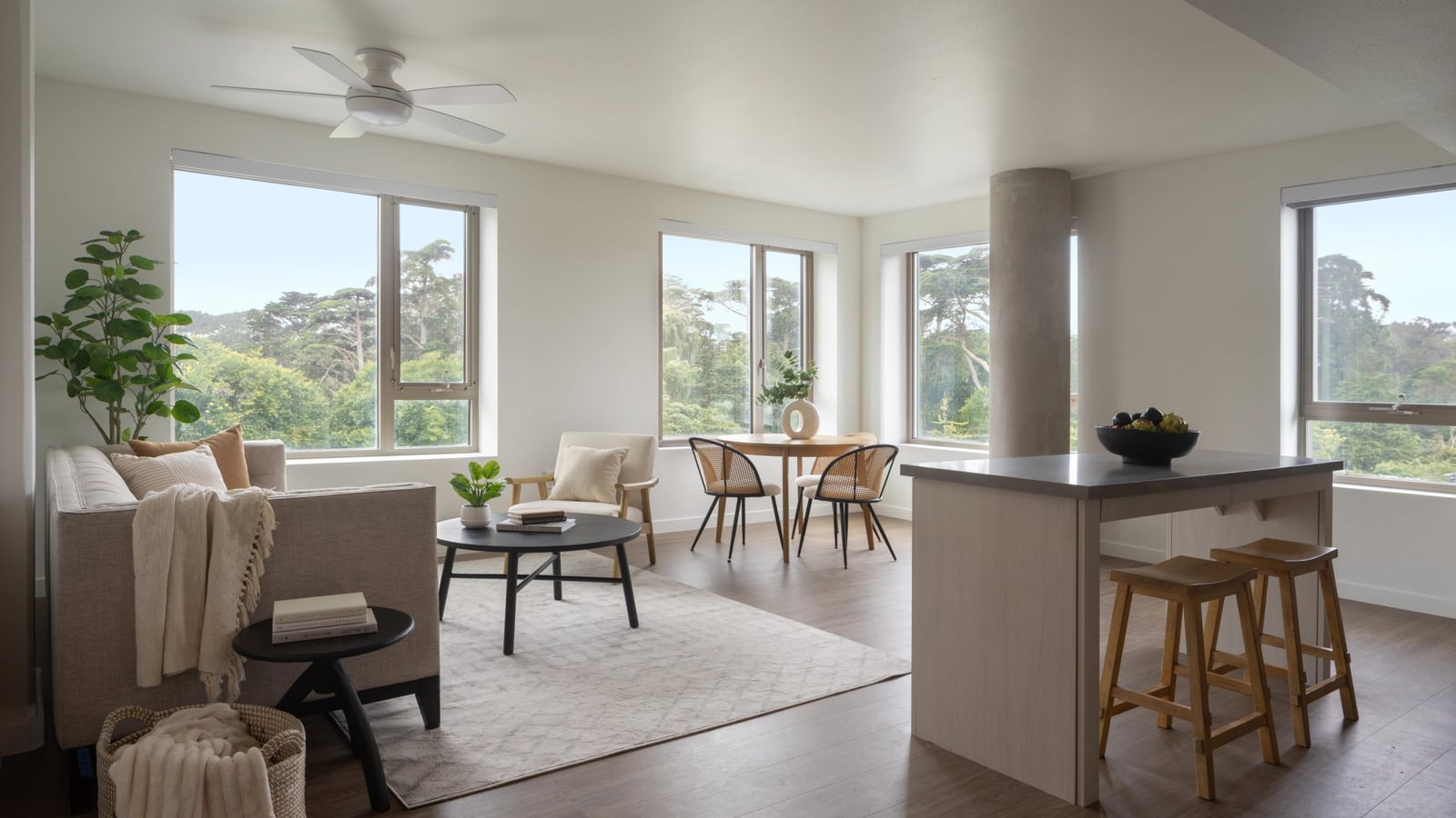 A furnished living and dining room with a sofa, a round dining table, a kitchen island with two stools, a ceiling fan and windows on two sides looking onto tall trees