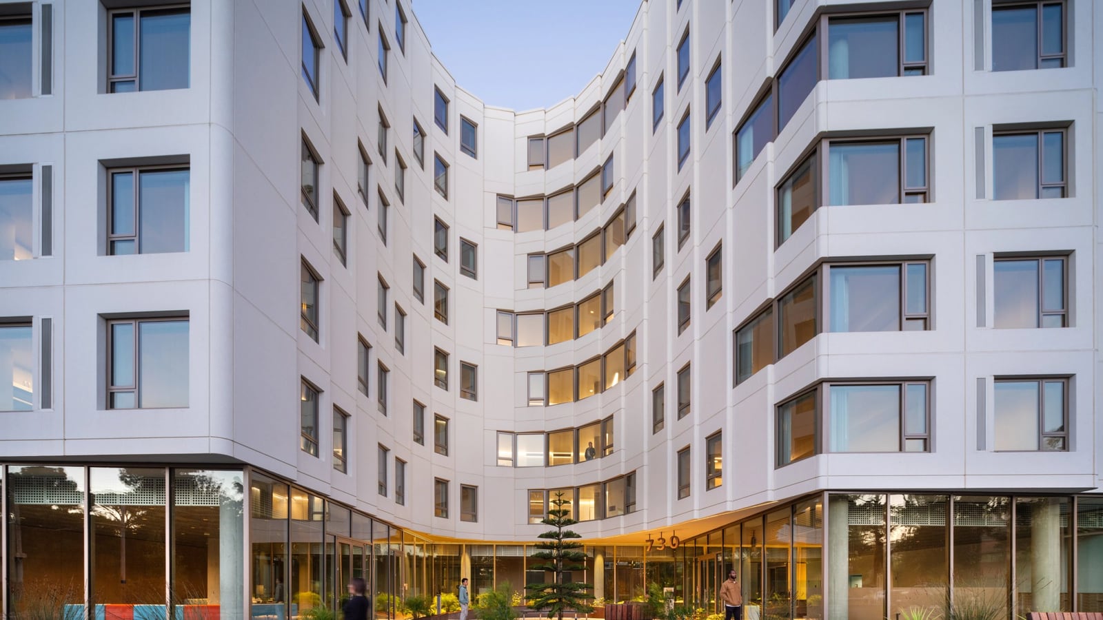 Curved white apartment wings with square windows rising above a glass-walled ground floor and a planted courtyard with benches