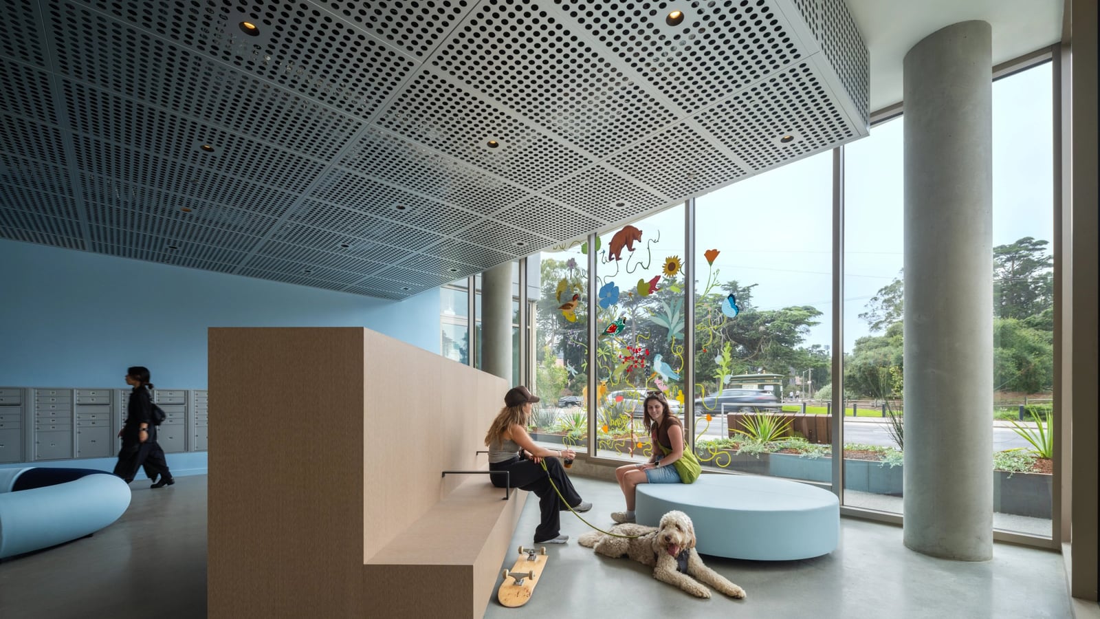 A lobby with a perforated metal ceiling, concrete columns, a wood bench where two people sit with a dog, mailboxes at left and floor-to-ceiling windows onto a street and park