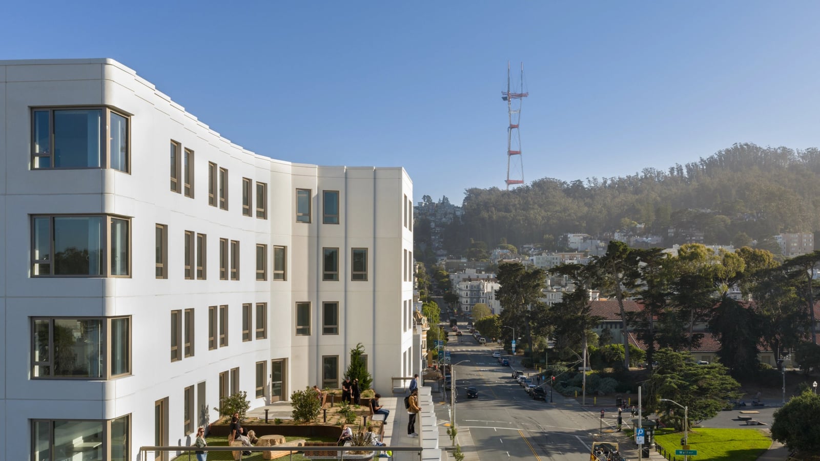 A curved white facade with rows of windows above a planted roof terrace with seating, with a street, a Muni bus, trees and Sutro Tower on the hill beyond