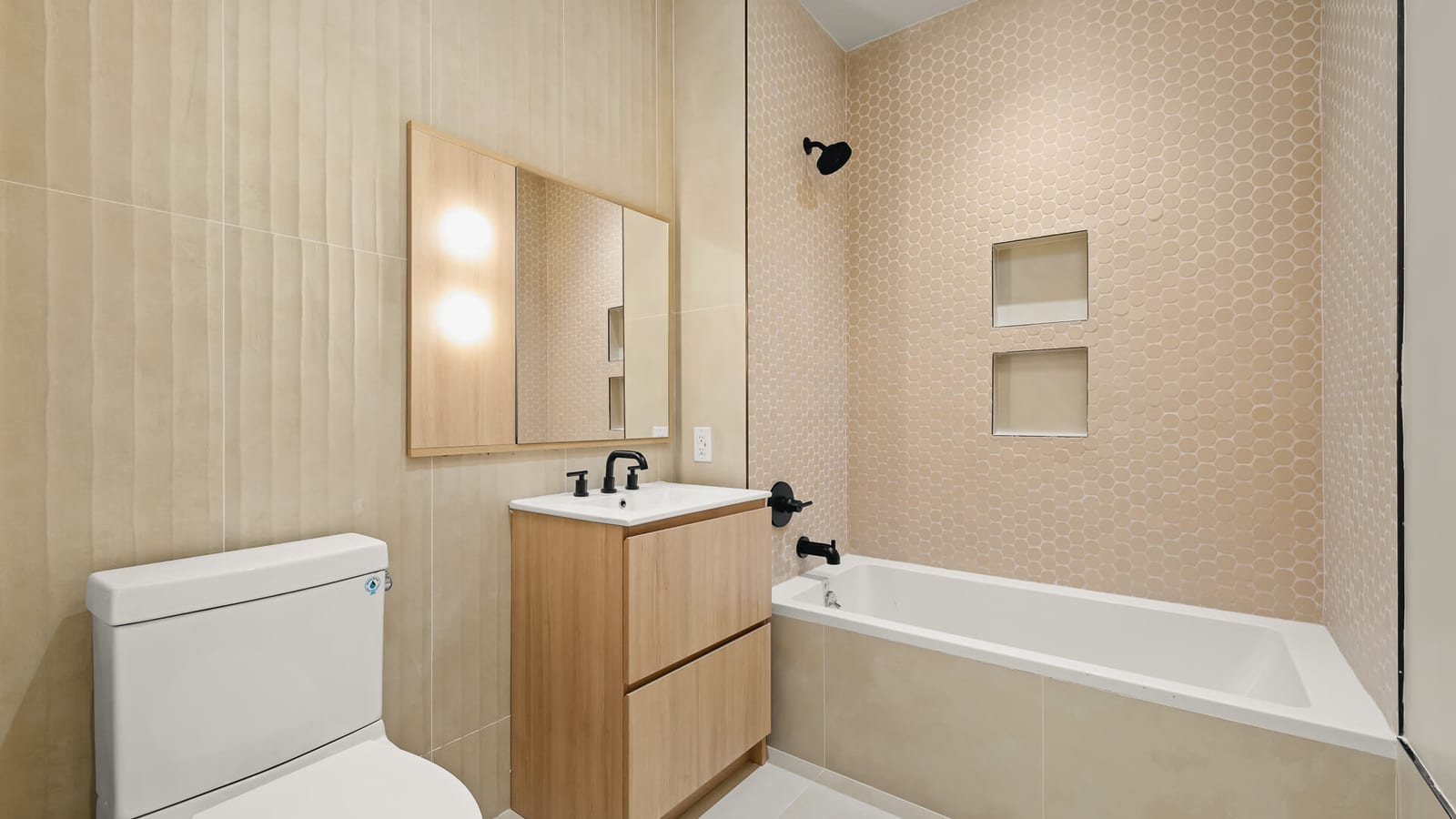 Photograph of a bathroom with beige ribbed wall tile, a light wood vanity with a white sink and black faucet, a lit mirror cabinet, a toilet, and a bathtub with pink penny-round tile, black fixtures and two wall niches