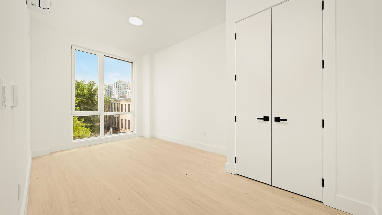 Photograph of an empty room with white walls, light wood-look floors, a white double closet, a wall-mounted air conditioning unit and a tall window looking onto brick row houses, trees and a newer tower