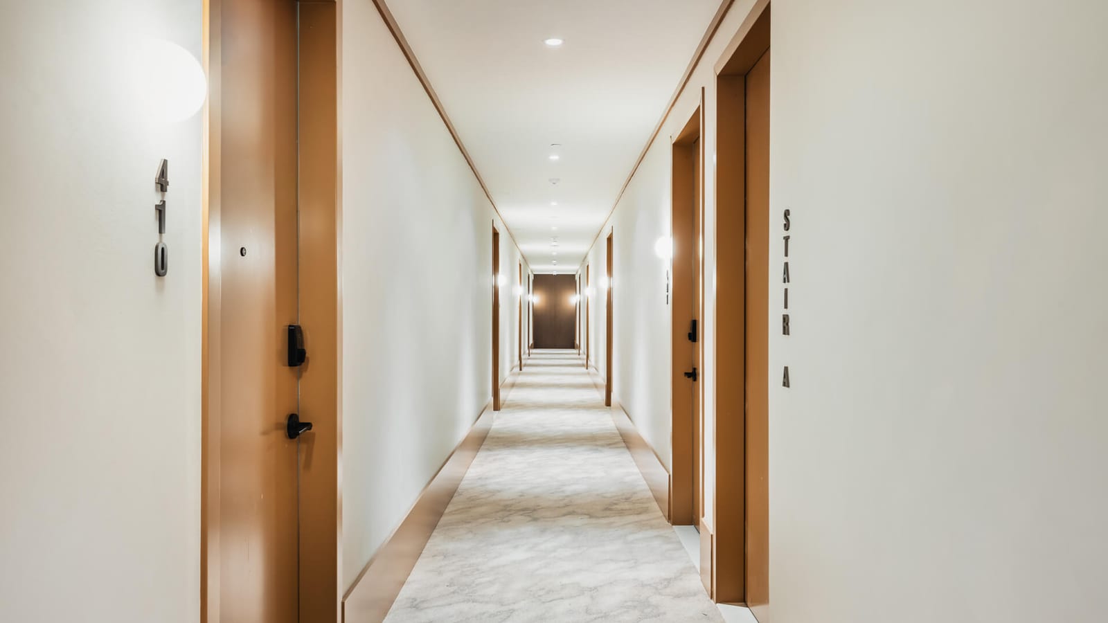 Photograph of a long hallway with white walls, wood-toned door frames and baseboards, a pale patterned carpet runner, wall lights, a door numbered 410 at left and a sign reading Stair A at right