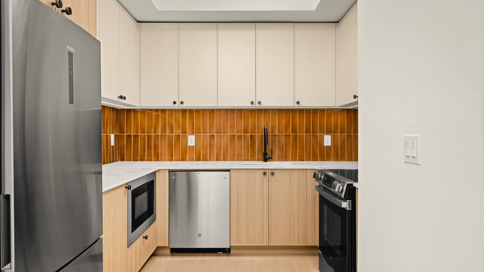 Photograph of a kitchen with cream upper cabinets, light wood lower cabinets, an amber tile backsplash, a white stone counter, a black faucet, a built-in microwave, a stainless dishwasher, a stainless refrigerator and a black and steel electric range