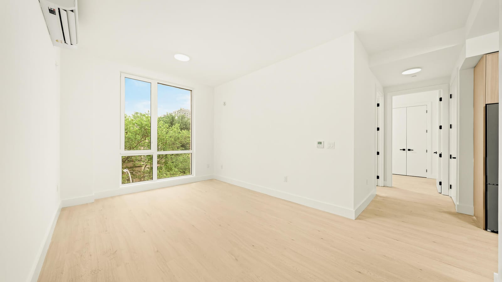 Photograph of an empty living room with white walls, light wood-look floors, a wall-mounted air conditioning unit, a tall two-part window looking onto trees and a tower, and a hall leading to closets and a dark refrigerator
