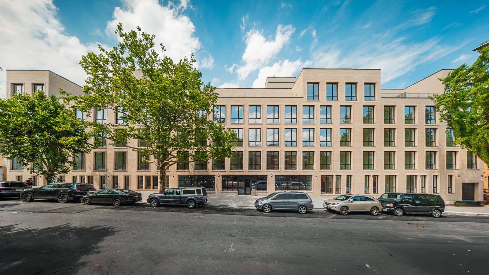 Photograph of a long five-story building of pale tan brick with rows of large dark-framed windows, a set-back top floor, a glass entrance at street level, street trees and parked cars in front, under a blue sky with white clouds