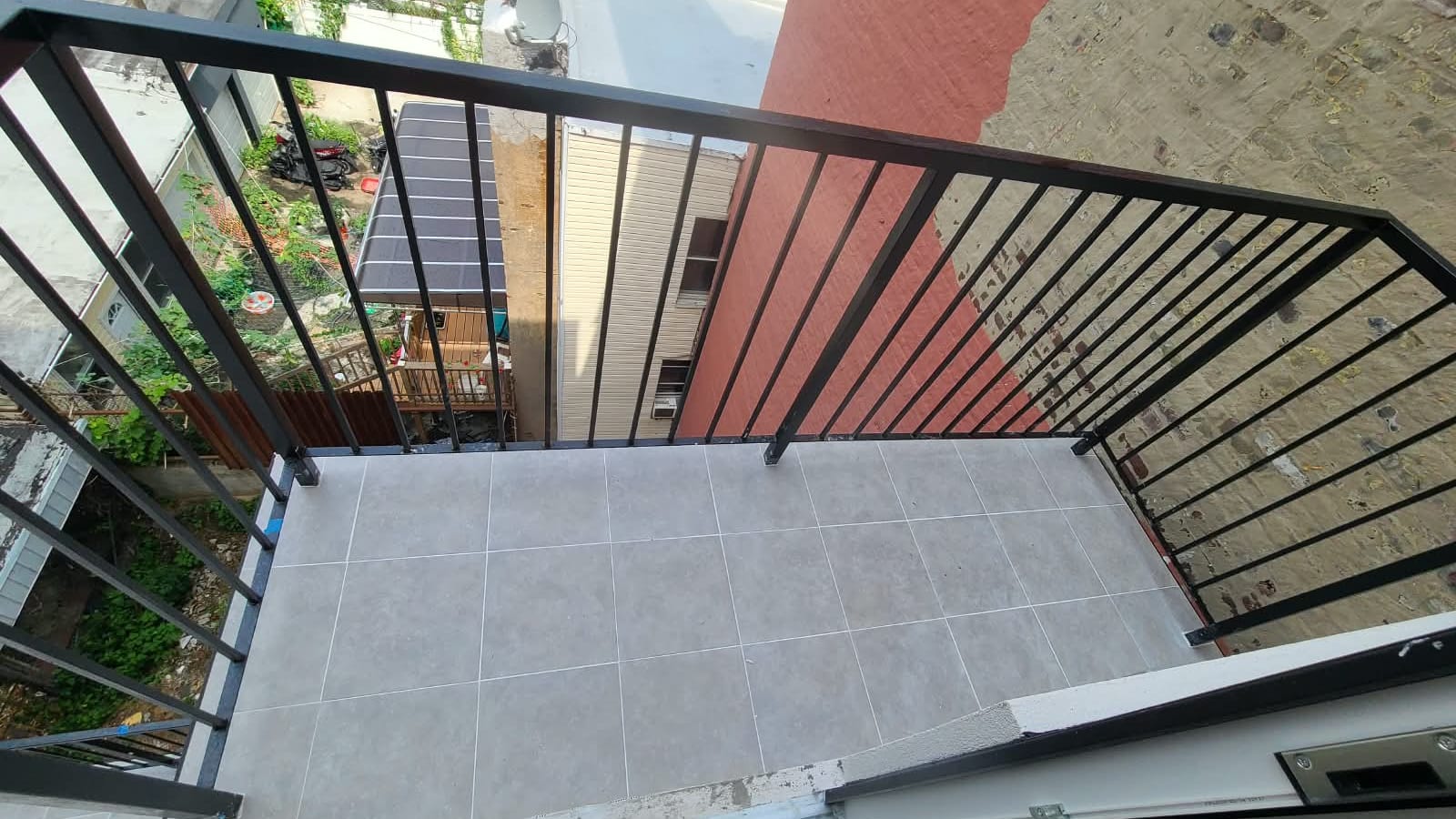 Photograph looking down onto a small balcony with grey square floor tiles and a black metal railing, beside a red-painted brick wall, with the backyards, fences and rooftops of neighboring houses below