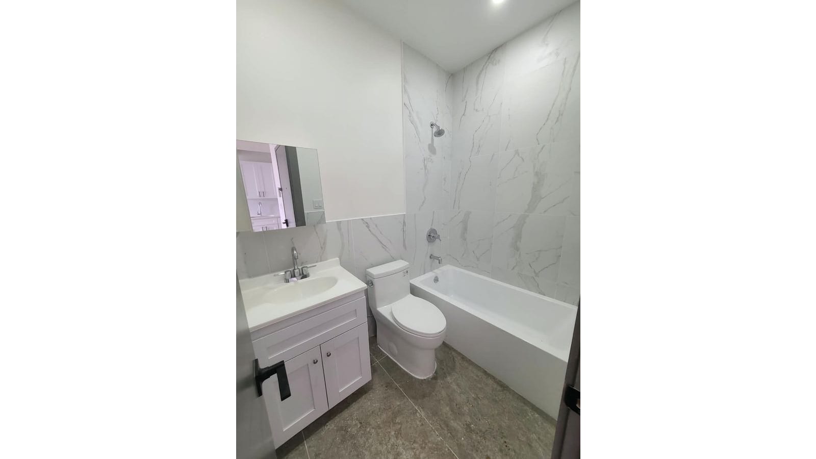 Photograph of a bathroom with white marble-look wall tiles, a bathtub with a chrome shower head, a white toilet, a white vanity sink cabinet, a wall mirror and a grey tile floor