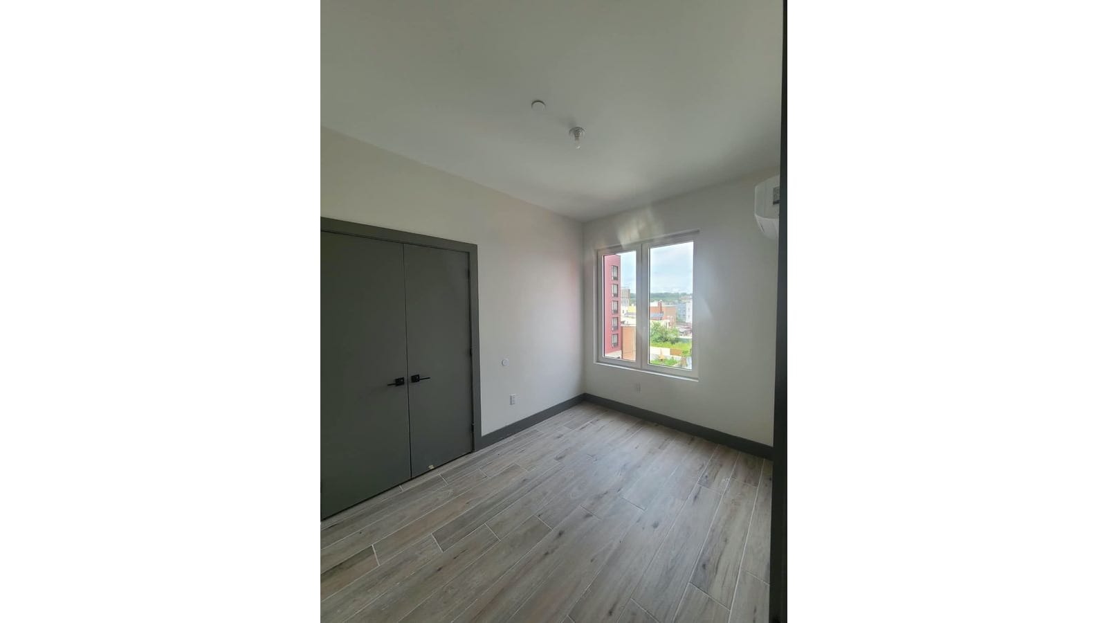 Photograph of an empty bedroom with wood-look tile floors, grey double closet doors, a white wall air-conditioning unit and a two-part window looking out over low buildings, trees and a taller red building