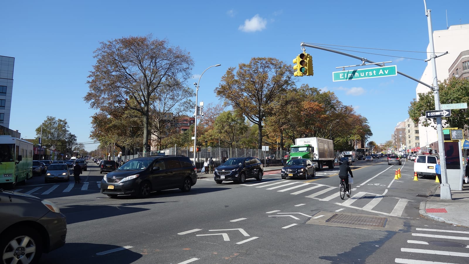 A wide street crossing under a clear blue sky, with traffic lights, a green Elmhurst Av street sign, cars, a green box truck and a cyclist seen from behind, and tall trees with autumn leaves behind a fence on the far side