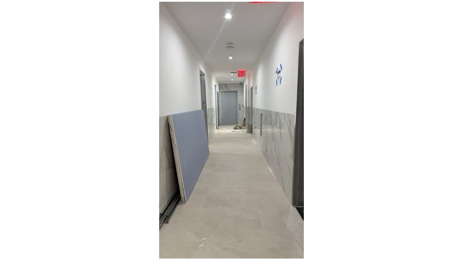A narrow building corridor with white upper walls and grey marble-look tile on the lower walls, doors on both sides, a lit red EXIT sign and, at the far end, a patterned metal door with a button panel beside it; a sheet of drywall leans on the left wall and cables lie on the floor