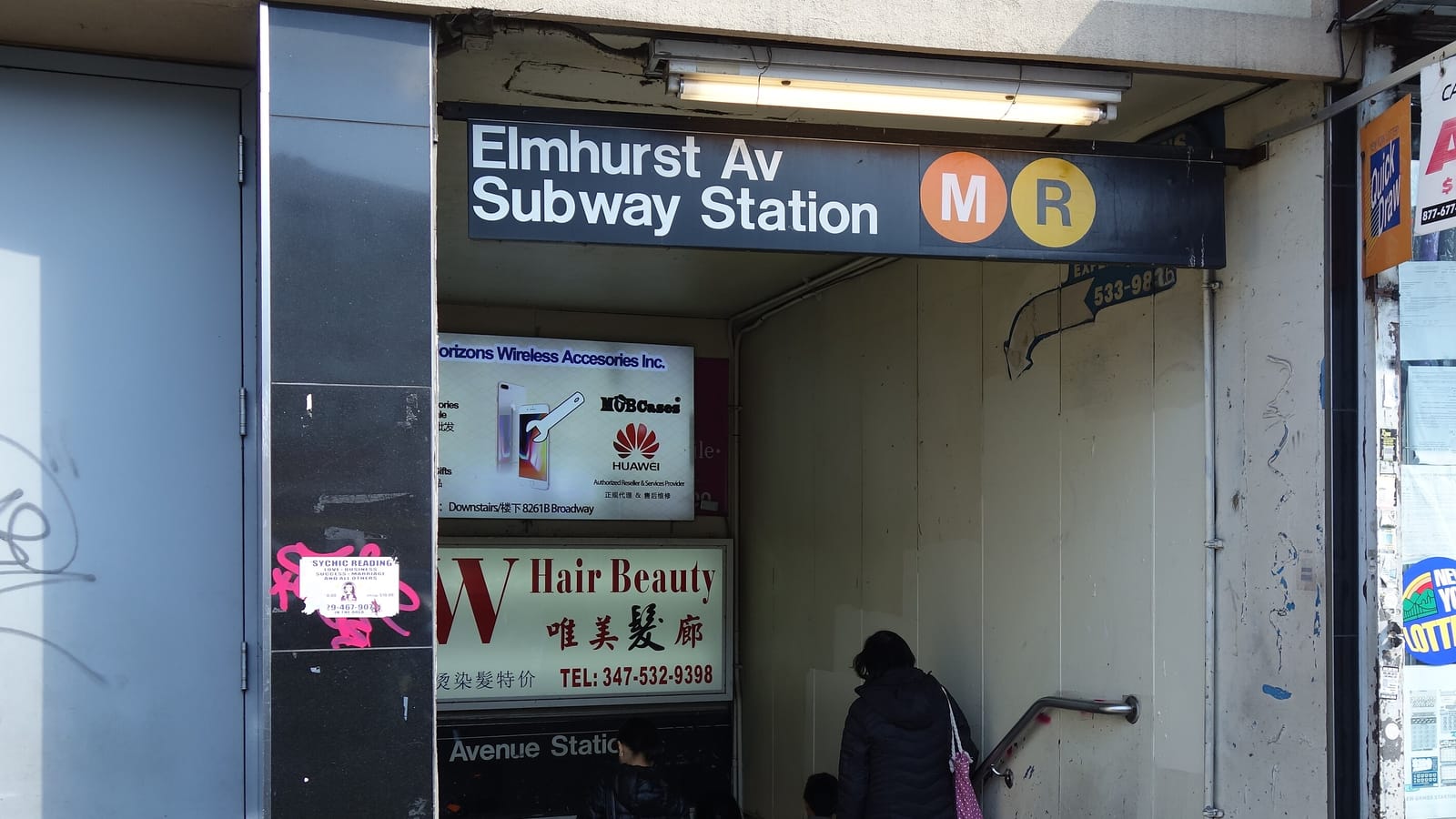 A subway entrance set into the front of a building, with a black sign reading Elmhurst Av Subway Station and orange M and yellow R circles over a stairway going down, shop signs on the walls inside and people seen from behind walking down the stairs