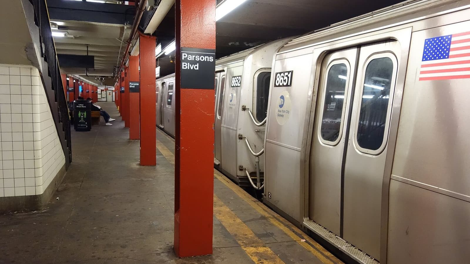 A subway platform with red columns, one carrying a black sign reading Parsons Blvd, beside a silver train with car numbers 8651 and 8652 and an American flag decal; benches and a trash can are far down the platform at left
