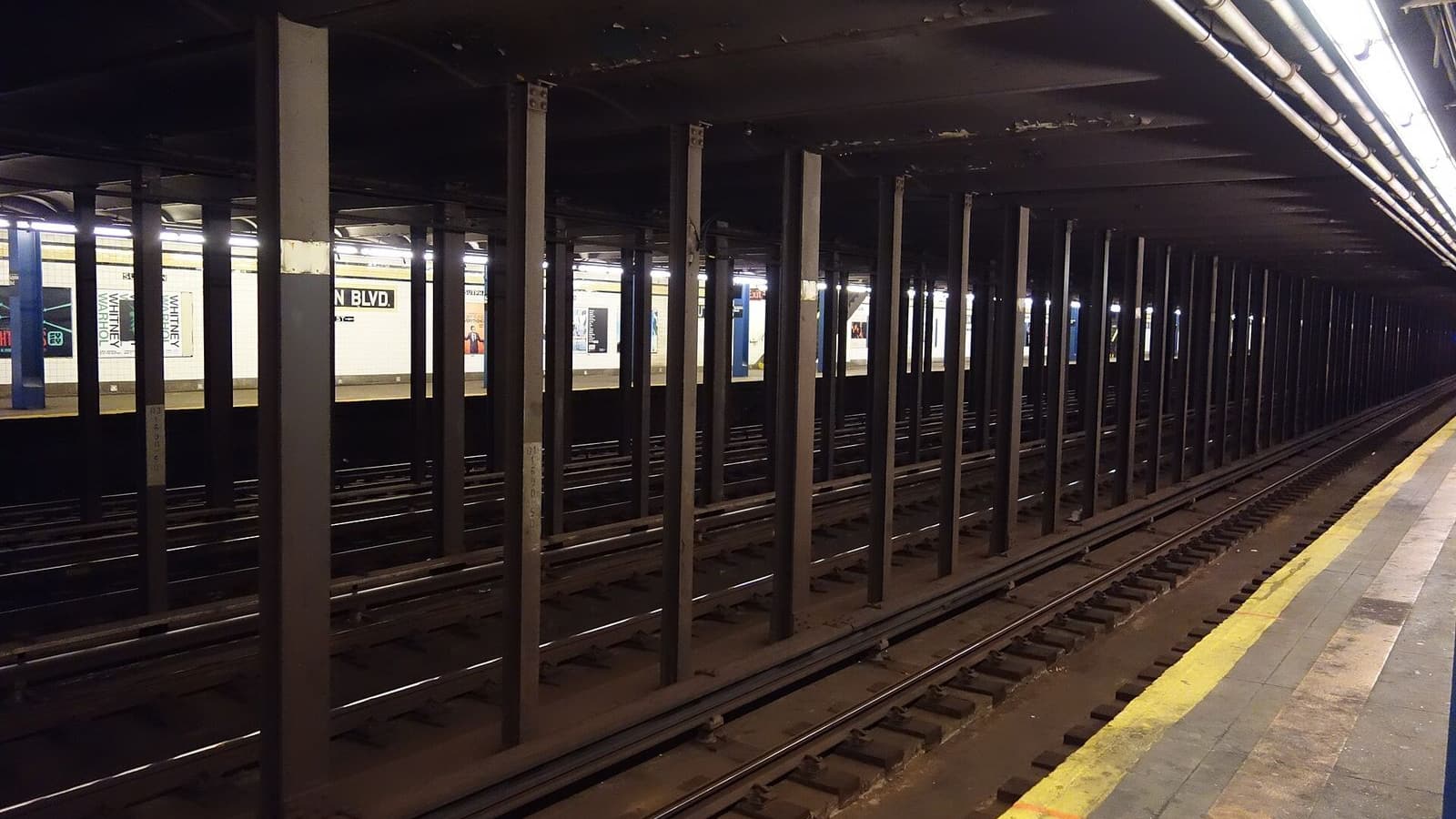 An empty subway platform edge with a yellow safety strip at right, several tracks separated by rows of dark steel columns, and the lit far platform with white tiles, a partly visible station name ending in BLVD and posters