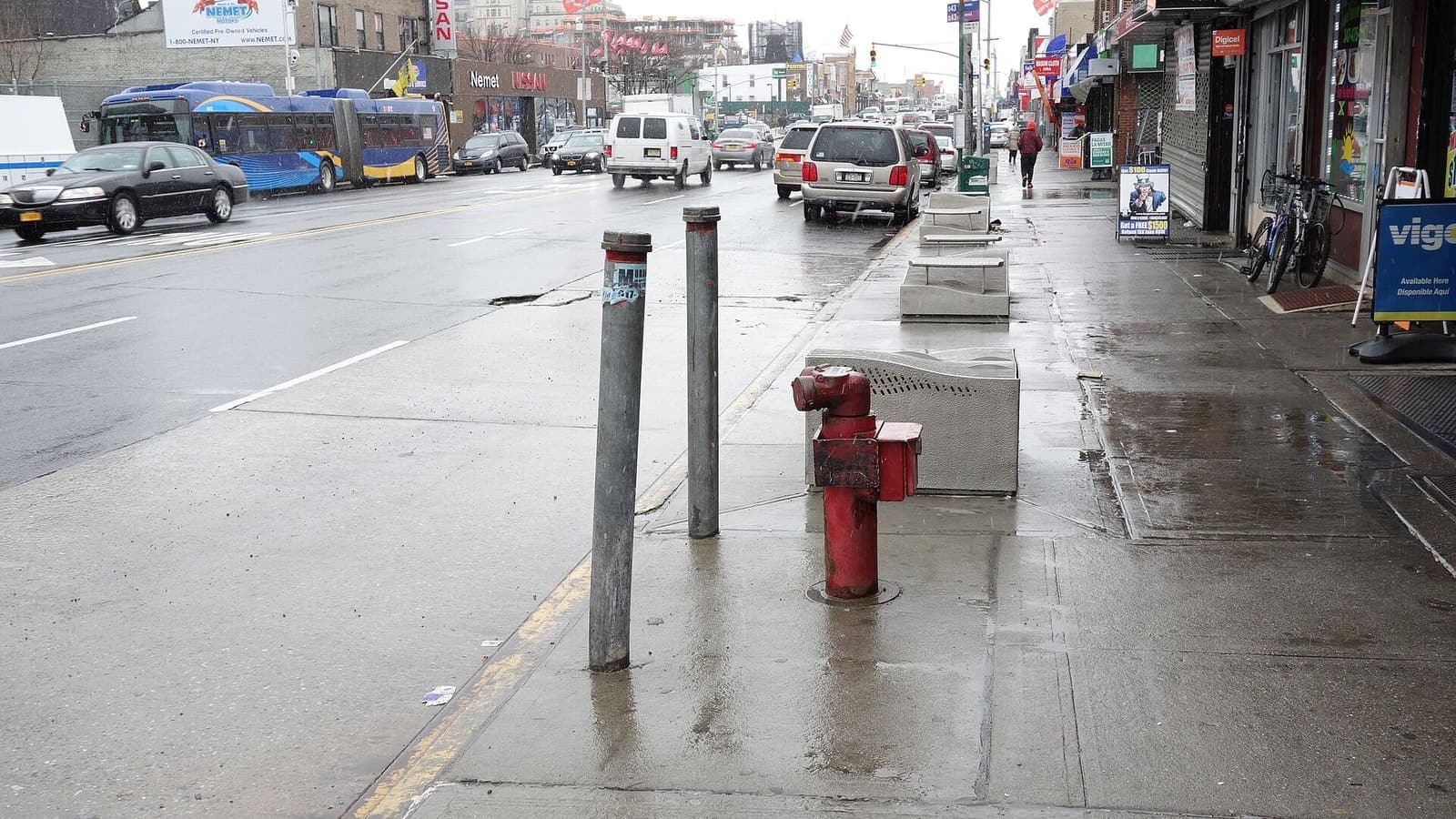 A wet sidewalk on Hillside Avenue with two metal posts, a red standpipe and concrete grates in front of storefronts at right; cars, a white van and a blue articulated bus drive along the avenue at left under a gray sky