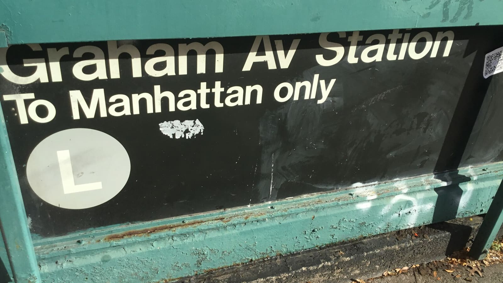 Photograph of a worn green subway entrance panel with a black sign reading Graham Av Station, To Manhattan only, and a white L train symbol, with fallen leaves on the sidewalk below
