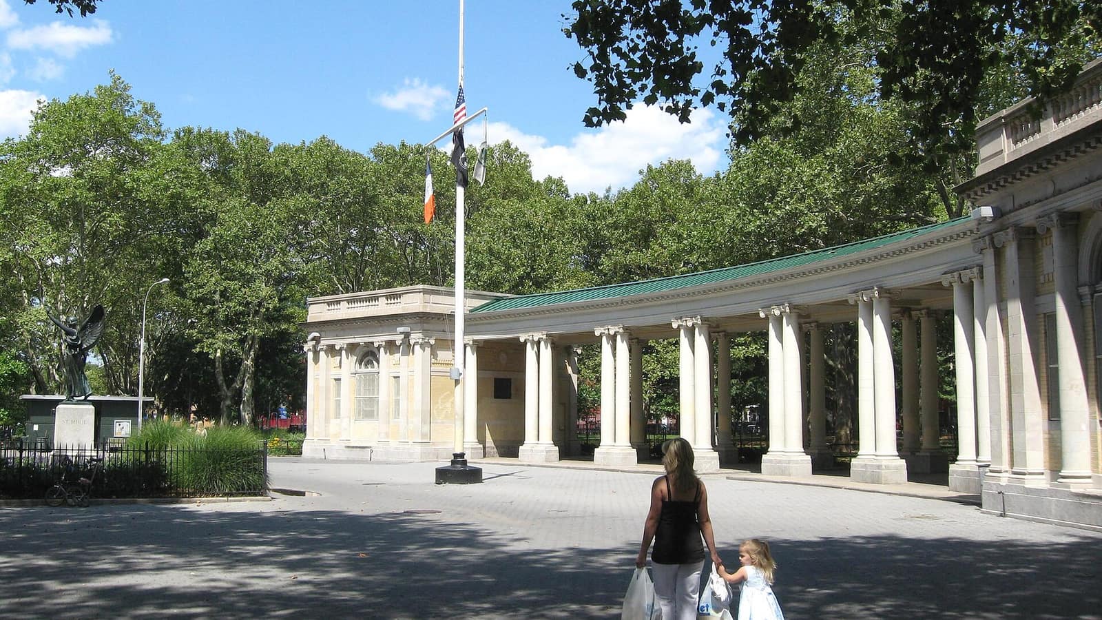 Photograph of a curved stone colonnade with a green roof and a small pavilion in a paved plaza, a flagpole with American and New York City flags, a winged bronze statue at left and tall trees, with a woman and a small child walking away from the camera