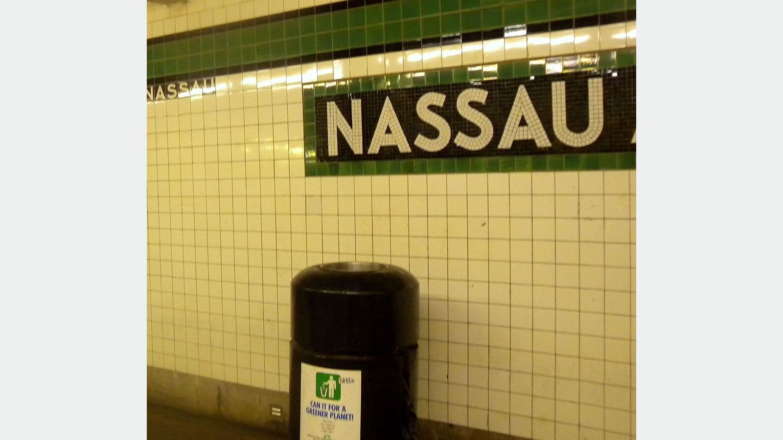 Photograph of a subway platform wall of white tiles with a green tile band and a black mosaic panel reading Nassau in white letters, and a black trash can with a recycling sticker, shown on a plain light background