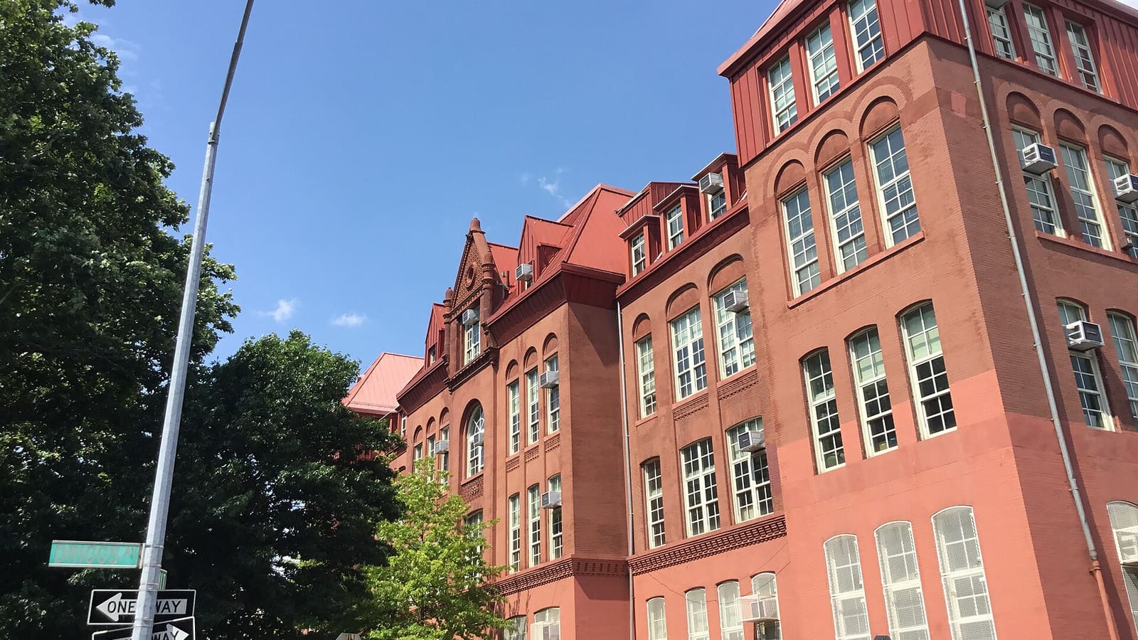 Photograph of a long four-story red brick school building with arched window bays, red metal roofs and dormers and window air conditioners, under a blue sky, with a street light, a green street sign and one-way signs at the corner and a large tree at left