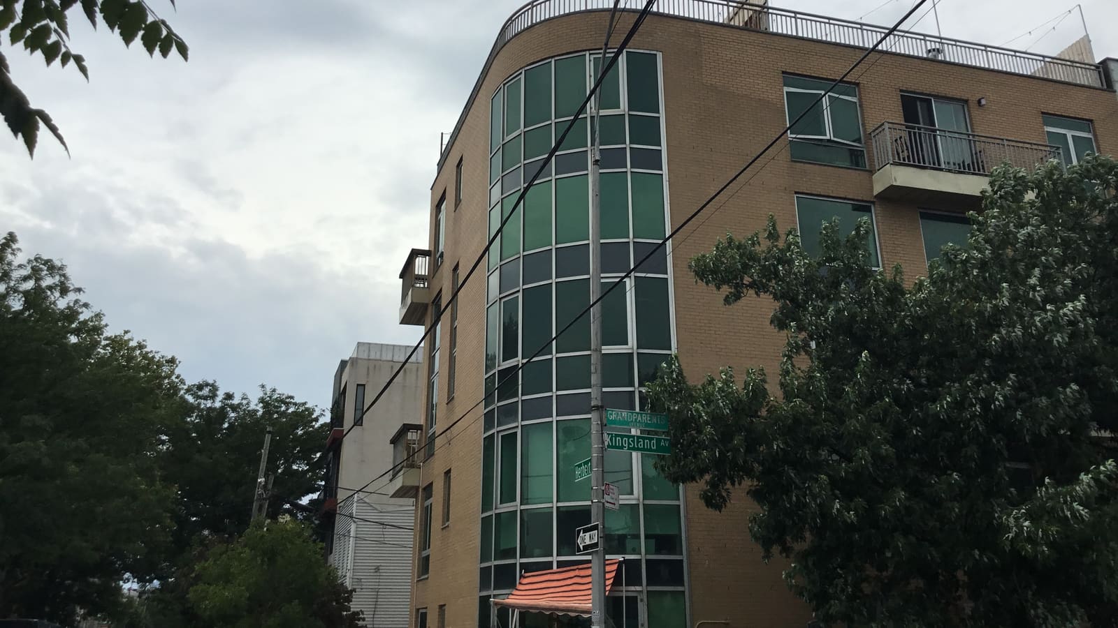 Photograph of a five-story tan brick corner building with a curved wall of green-tinted windows, a rooftop railing and small balconies, under a cloudy sky, with a utility pole carrying green street signs reading Grandparents Avenue, Kingsland Av and Herbert, a one-way sign, a striped awning at street level and leafy trees on both sides