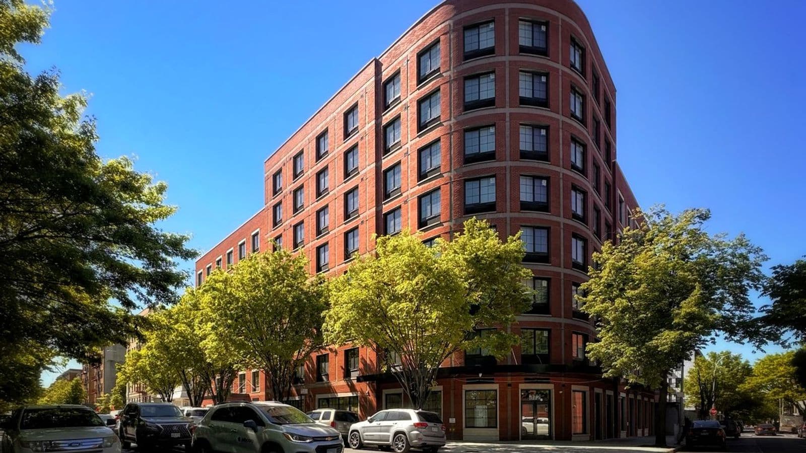Photograph of an eight-story red brick apartment building with a rounded corner, rows of black-framed windows set in light gray trim, a black metal canopy over the ground floor, street trees in leaf and parked cars at the curb under a blue sky