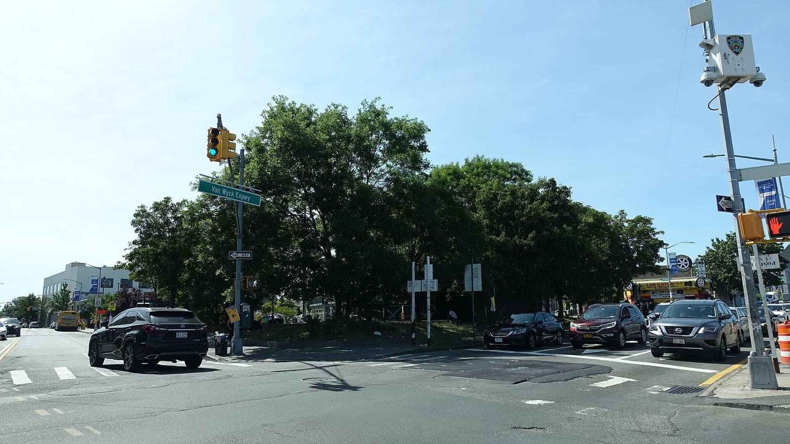 Photograph of a wide street intersection on a sunny day with a traffic light and a green street sign reading Van Wyck Expwy, a triangle of tall trees behind a low fence, a line of cars waiting at the crosswalk, a pole with a police camera at right and low commercial buildings in the distance