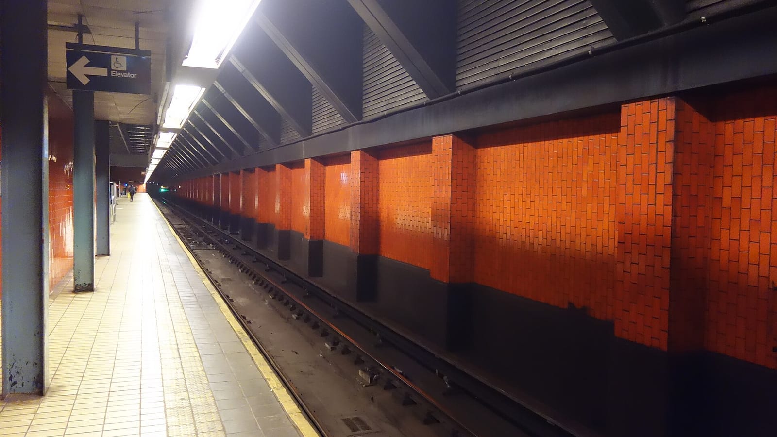 Photograph of an underground subway platform with orange-red tile walls along the tracks, dark steel beams overhead, a row of ceiling lights, a hanging sign with a wheelchair symbol reading Elevator with an arrow to the left, and one small distant figure at the far end of the platform
