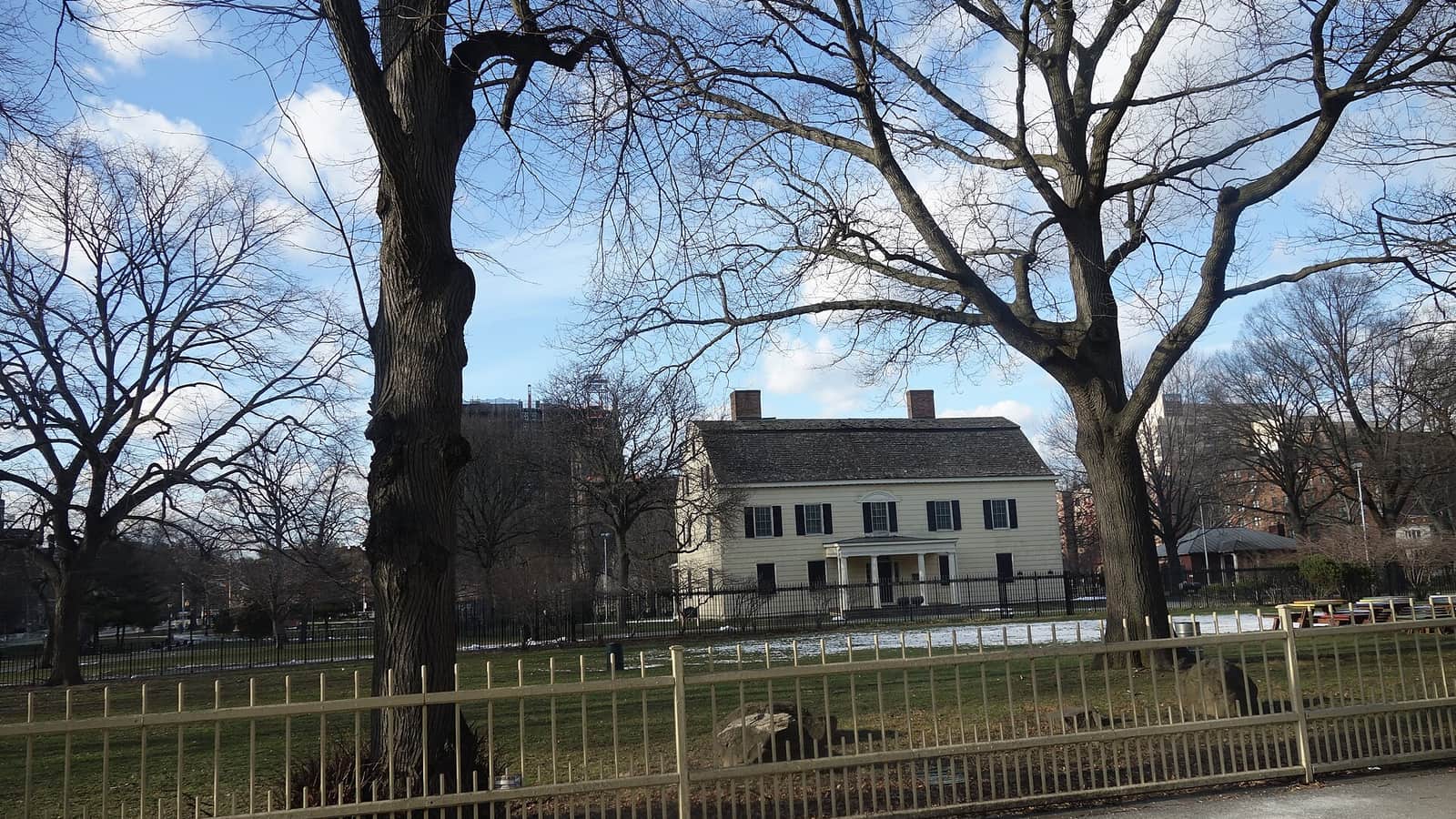 Photograph of a two-story pale yellow clapboard house with black shutters, two chimneys and a small columned porch, standing on a lawn with patches of snow behind a black iron fence, seen through bare winter trees and a cream-colored metal fence in the foreground under a blue sky with clouds