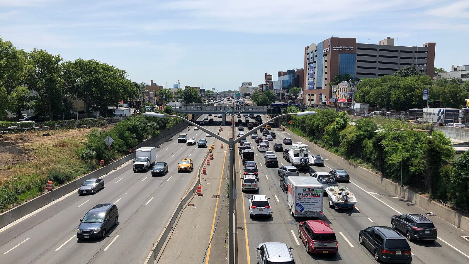 Photograph looking down on a sunken eight-lane expressway full of cars, a box truck and a car towing a boat, with a double street lamp in the median, an overpass in the distance, trees along both sides and a tall brick and glass building with a sign reading Jamaica Hospital Medical Center on the right under a pale blue sky