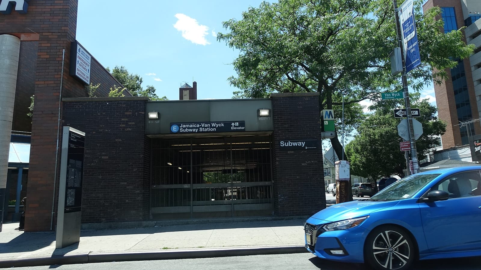 Photograph of a dark brick subway entrance on a sunny day with a sign reading E Jamaica-Van Wyck Subway Station and an arrow to the elevator, a Subway sign and a green M elevator marker on the right, a green Van Wyck Expressway street sign on a pole, a tree behind, a tall brick hospital building with an Emergency Entrance sign at left and a blue car passing in front
